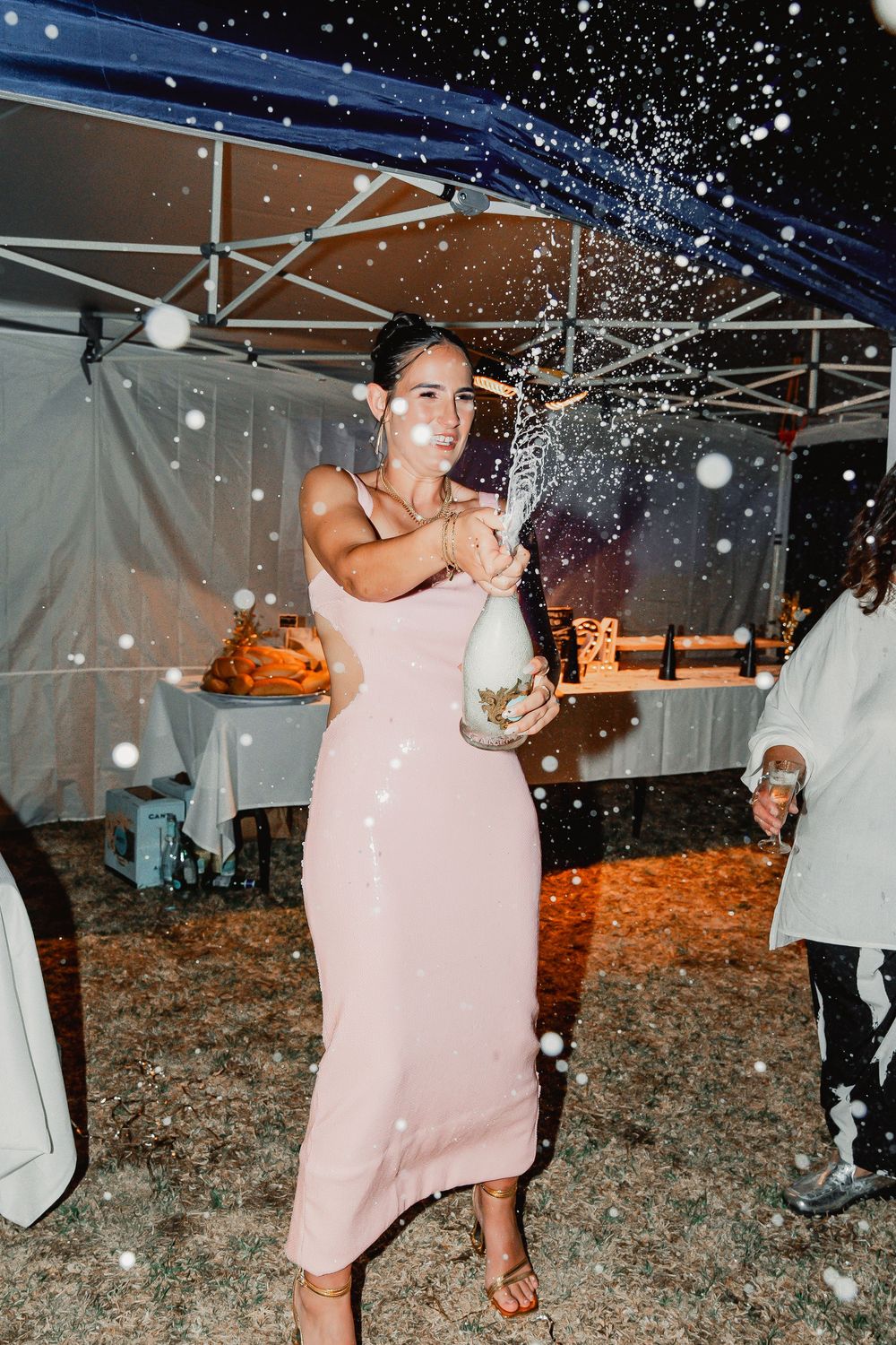 Someone in a pink fitted dress sprays champagne during a celebration under an outdoor tent while partygoers look on.