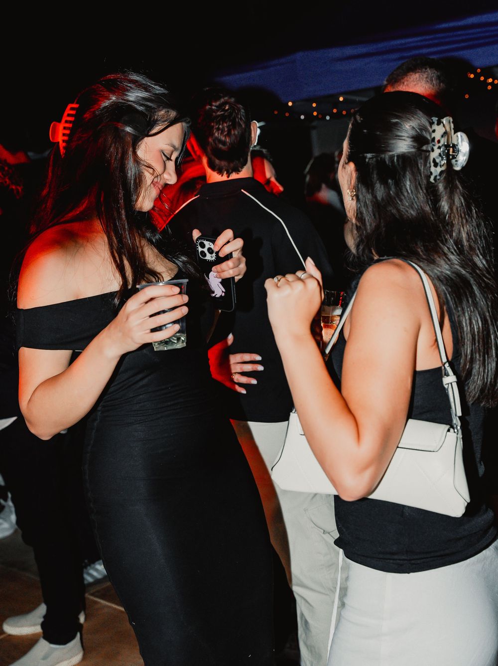Two friends in black and white outfits sharing drinks and laughing at a party with red lighting.