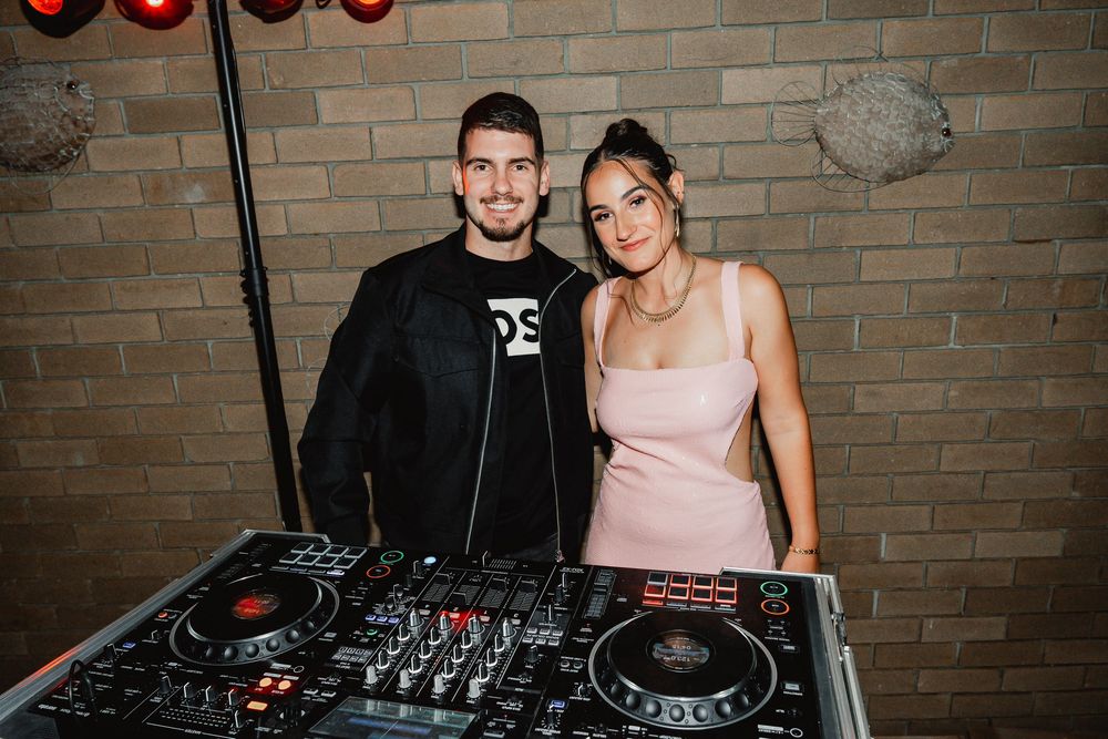 Two DJs stand behind professional DJ equipment with a brick wall background in a nightclub setting.