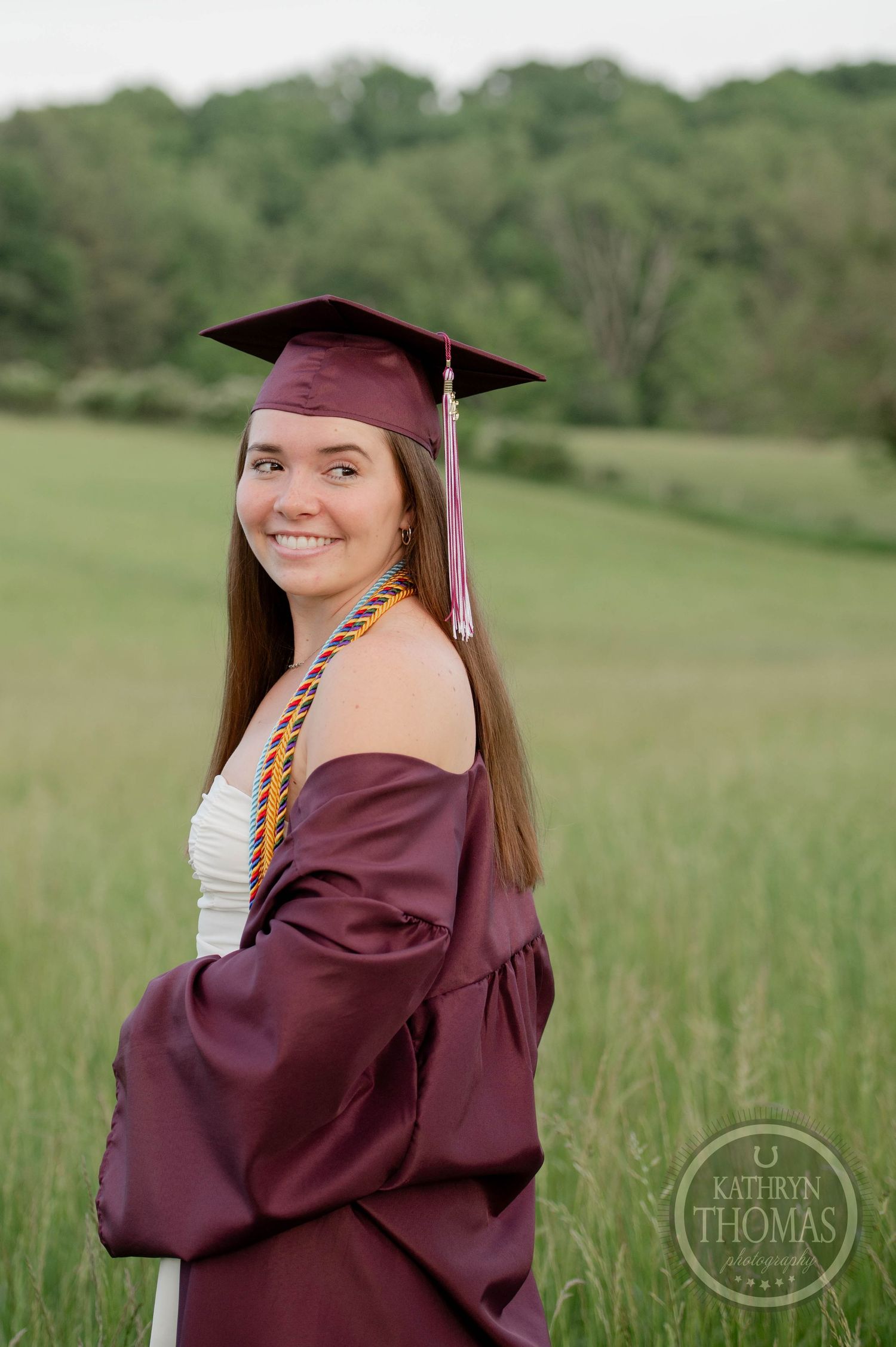 Graduate in burgundy cap and gown poses in sunny meadow wearing honor cords and white dress underneath.