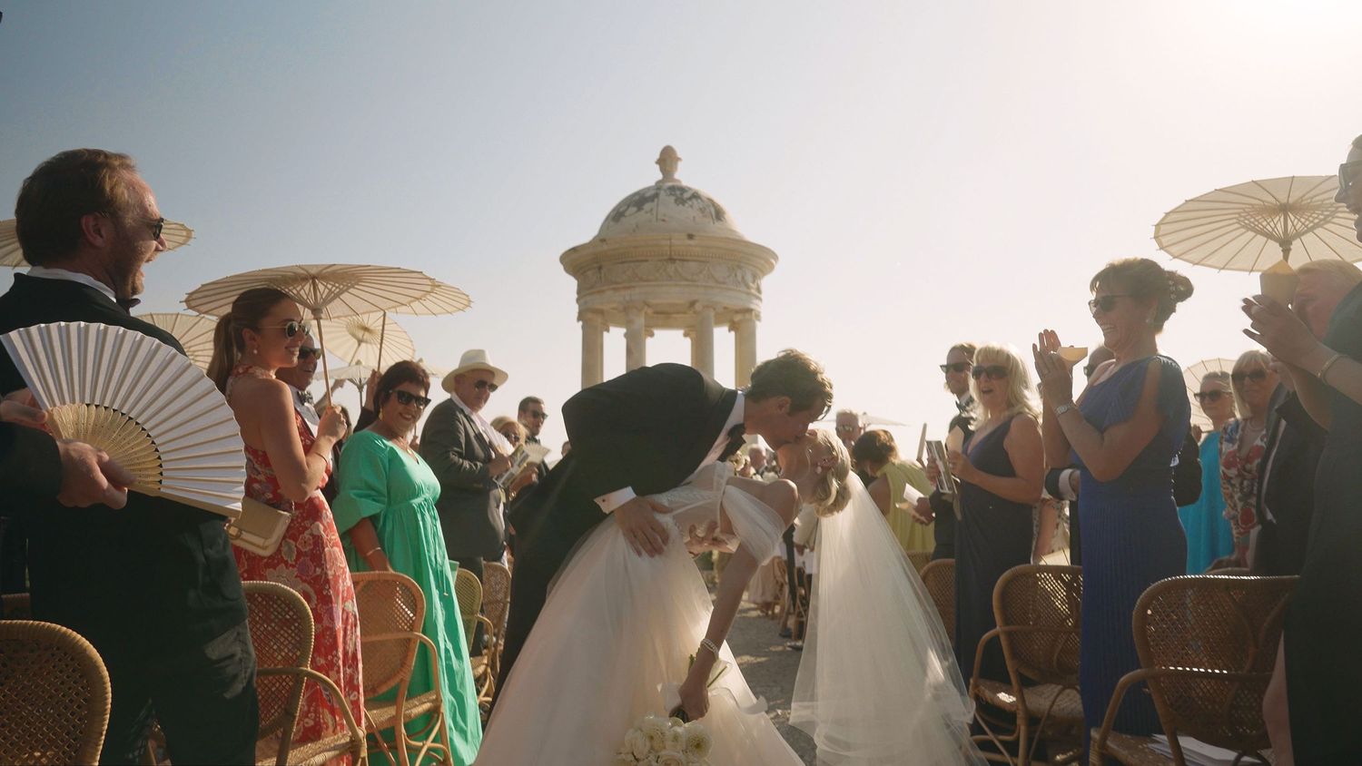 Brides sharing a kiss at their outdoor wedding, surrounded by guests celebrating under the sun.