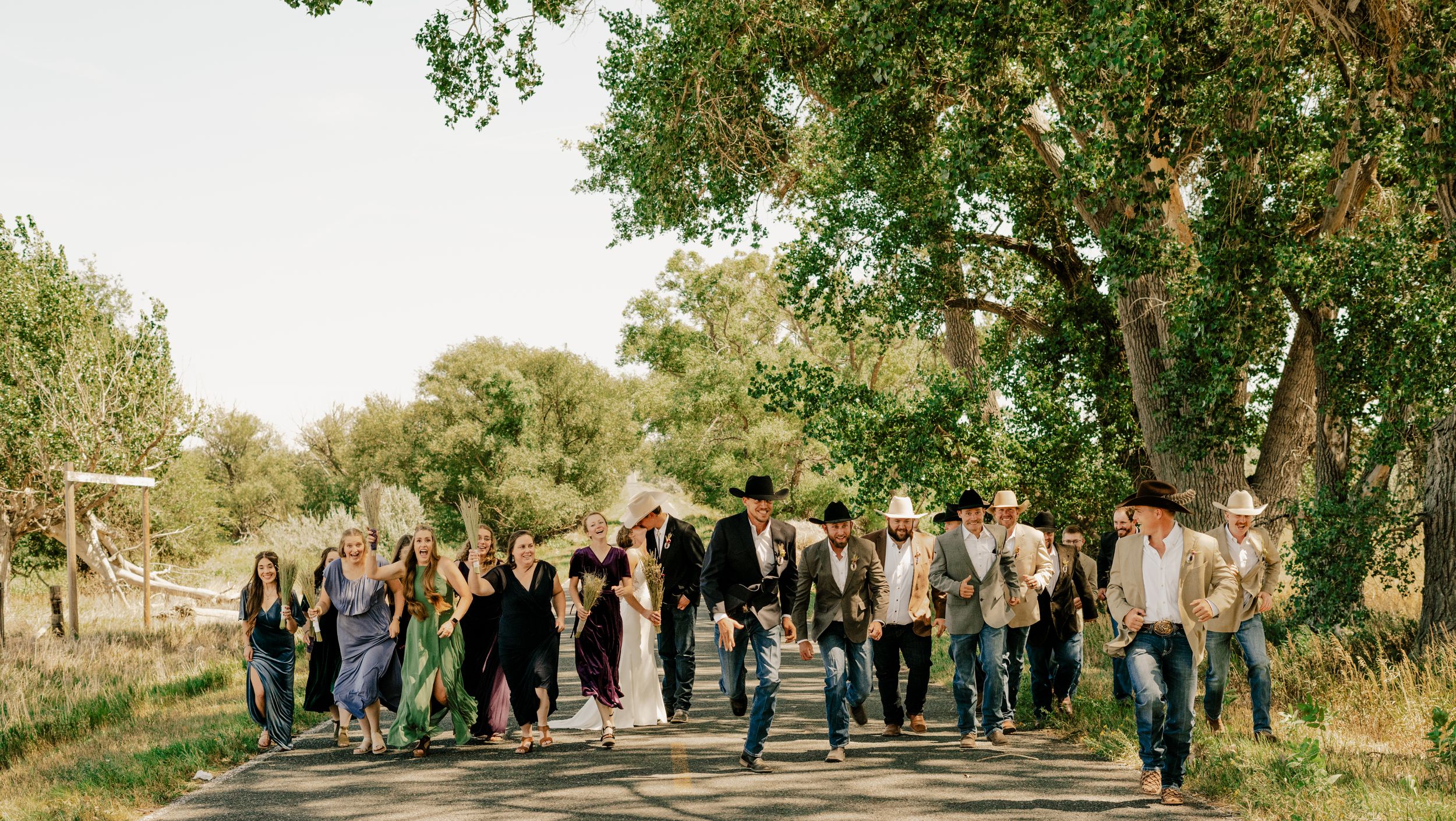 A bridal party running towards the camera with the bride and groom kissing in the background
