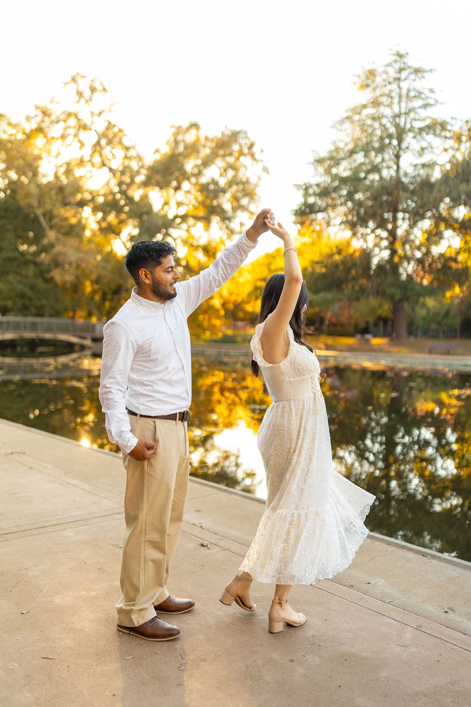 Couple in white and beige outfits dancing together by a pond during golden hour.