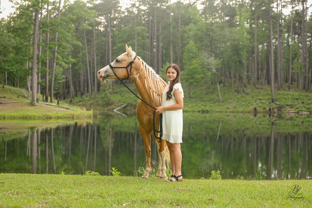 A young woman in a white dress stands beside a golden palomino horse near a serene pond surrounded by lush green trees.