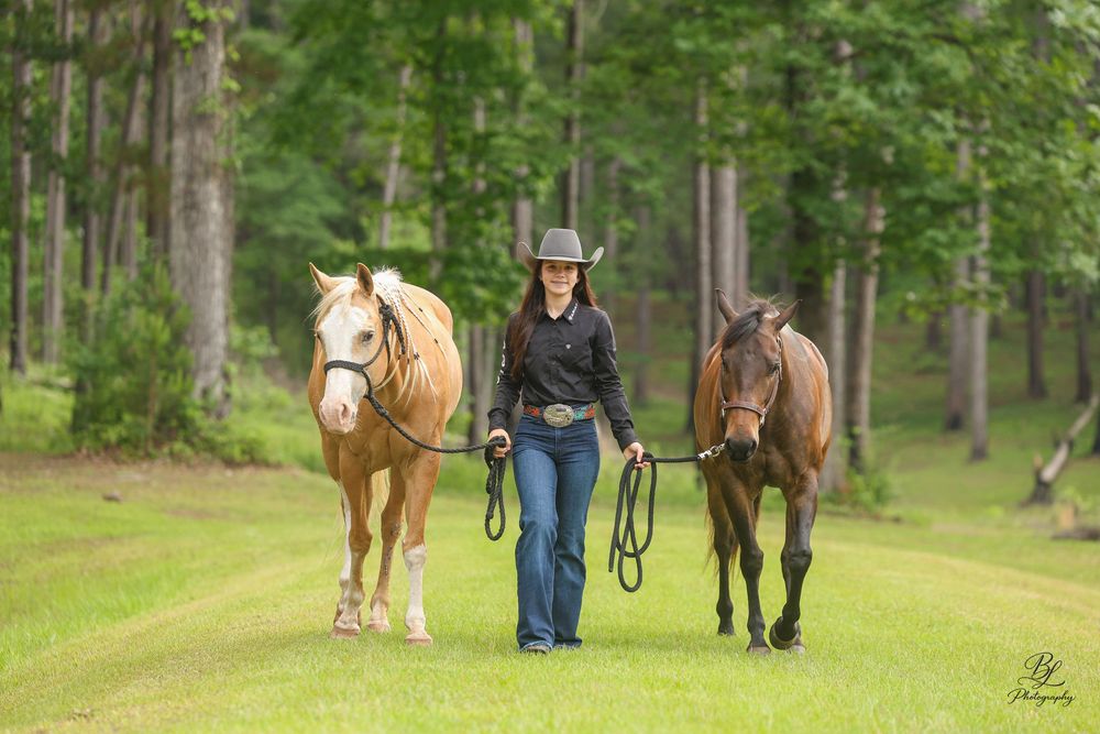Woman in cowboy hat walks two horses down a grassy path surrounded by lush green trees.