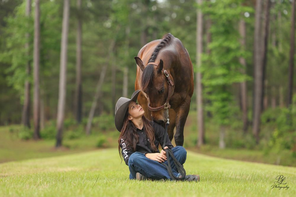 Woman in cowboy hat sits on grass as a brown horse leans down to nuzzle her face.