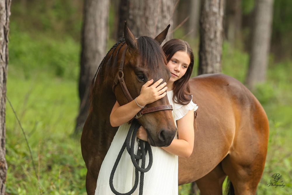 A young girl in a white dress tenderly embraces a brown horse in a lush green forest setting.