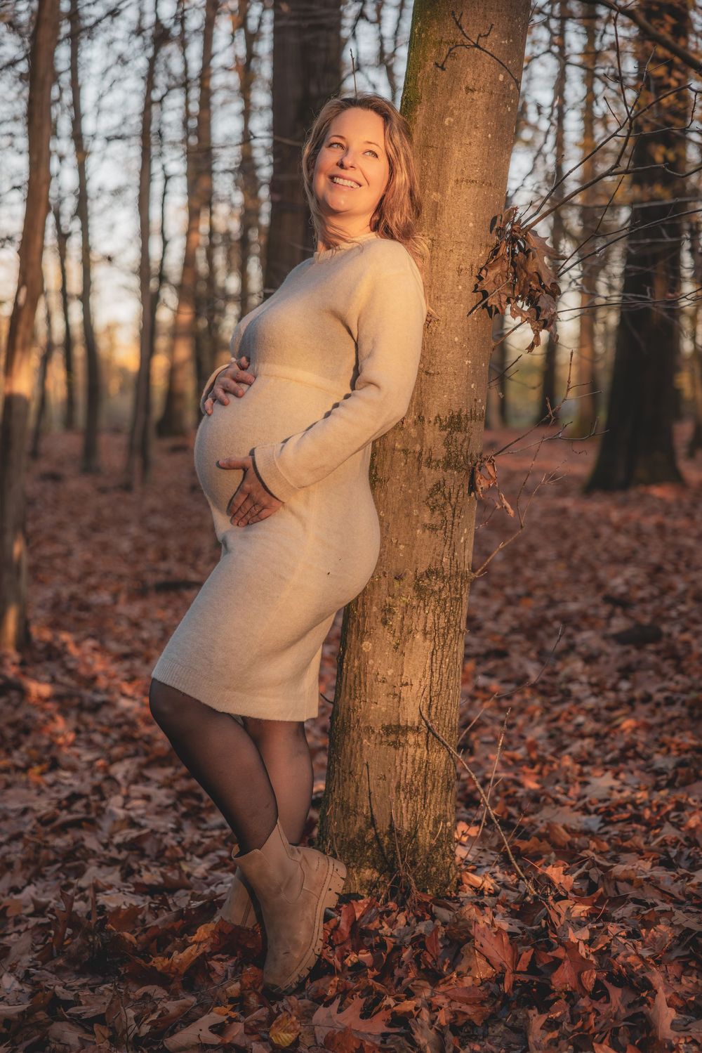 Person in cream sweater poses playfully against tree trunk in autumn forest with fallen orange leaves.
