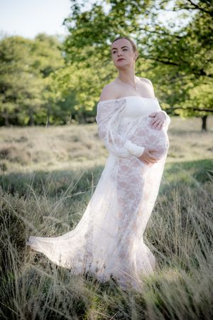 Person wearing an off-shoulder white lace gown poses elegantly in a sunlit field with trees in the background.