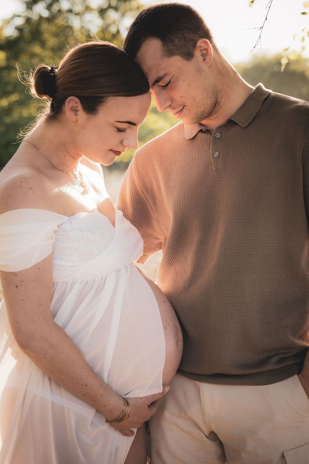 A couple shares an intimate moment during a maternity photoshoot at golden hour outdoors.