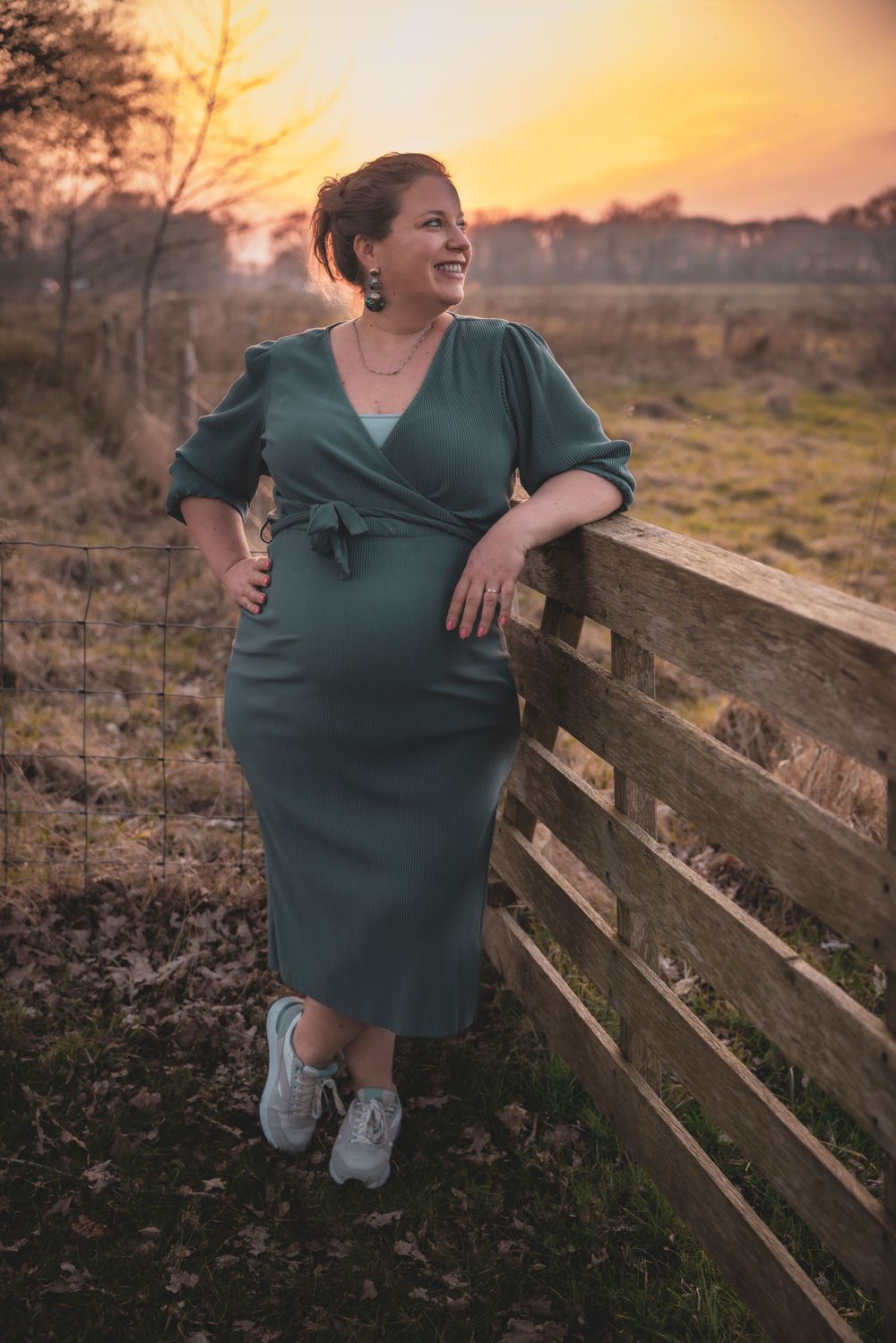 A figure in a green wrap dress and sneakers poses against a wooden fence at sunset in a rural setting.
