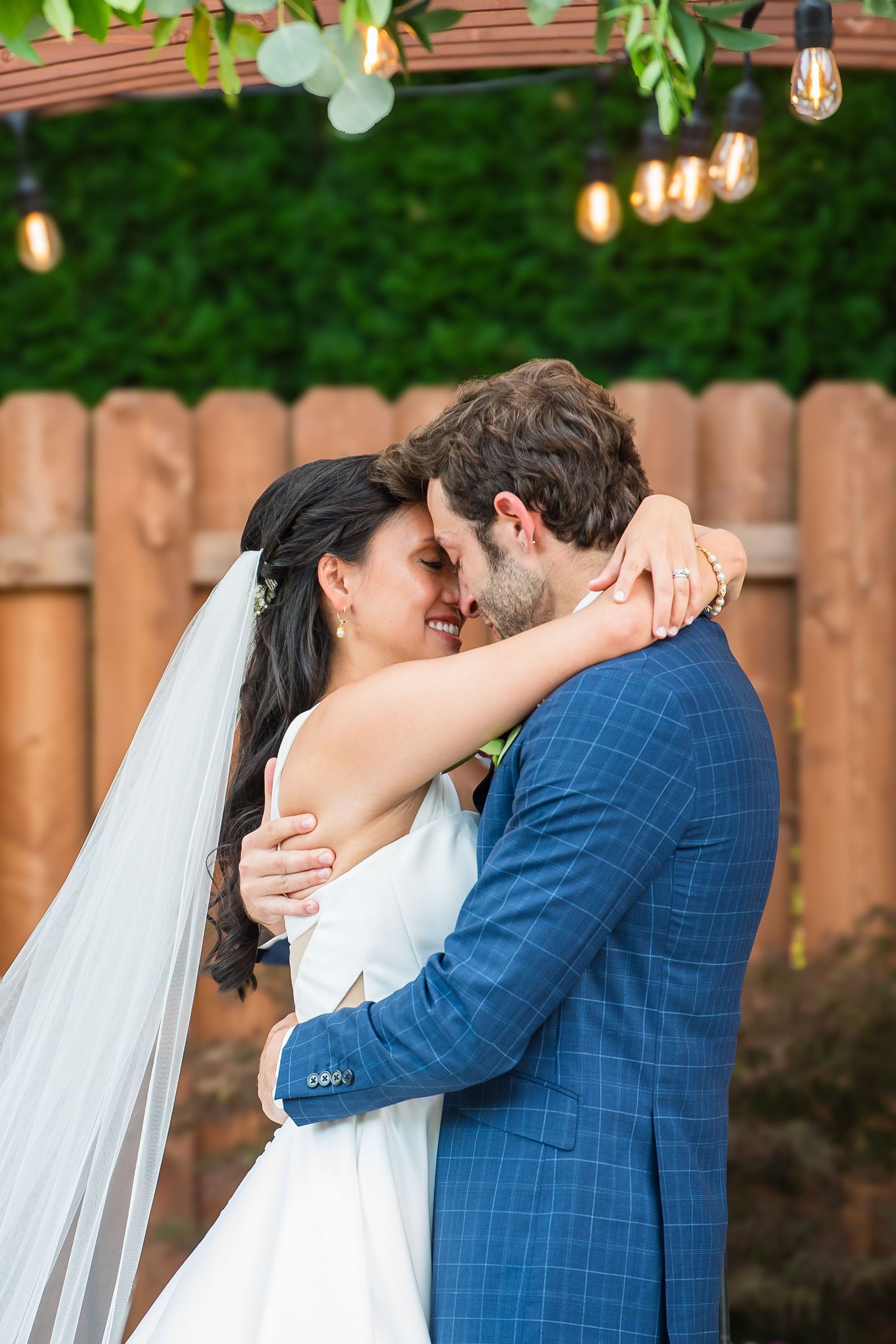Bride and groom share a romantic embrace outdoors, with string lights and lush greenery backdrop at their wedding.