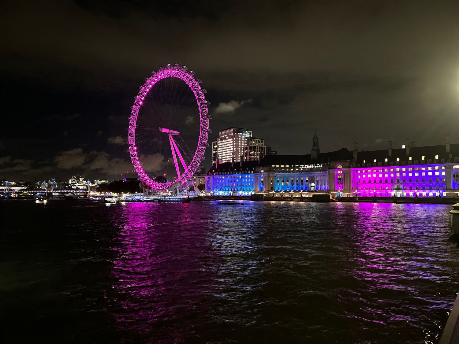 London Eye Ferris wheel illuminated in pink and purple lights reflecting on the Thames River at night.