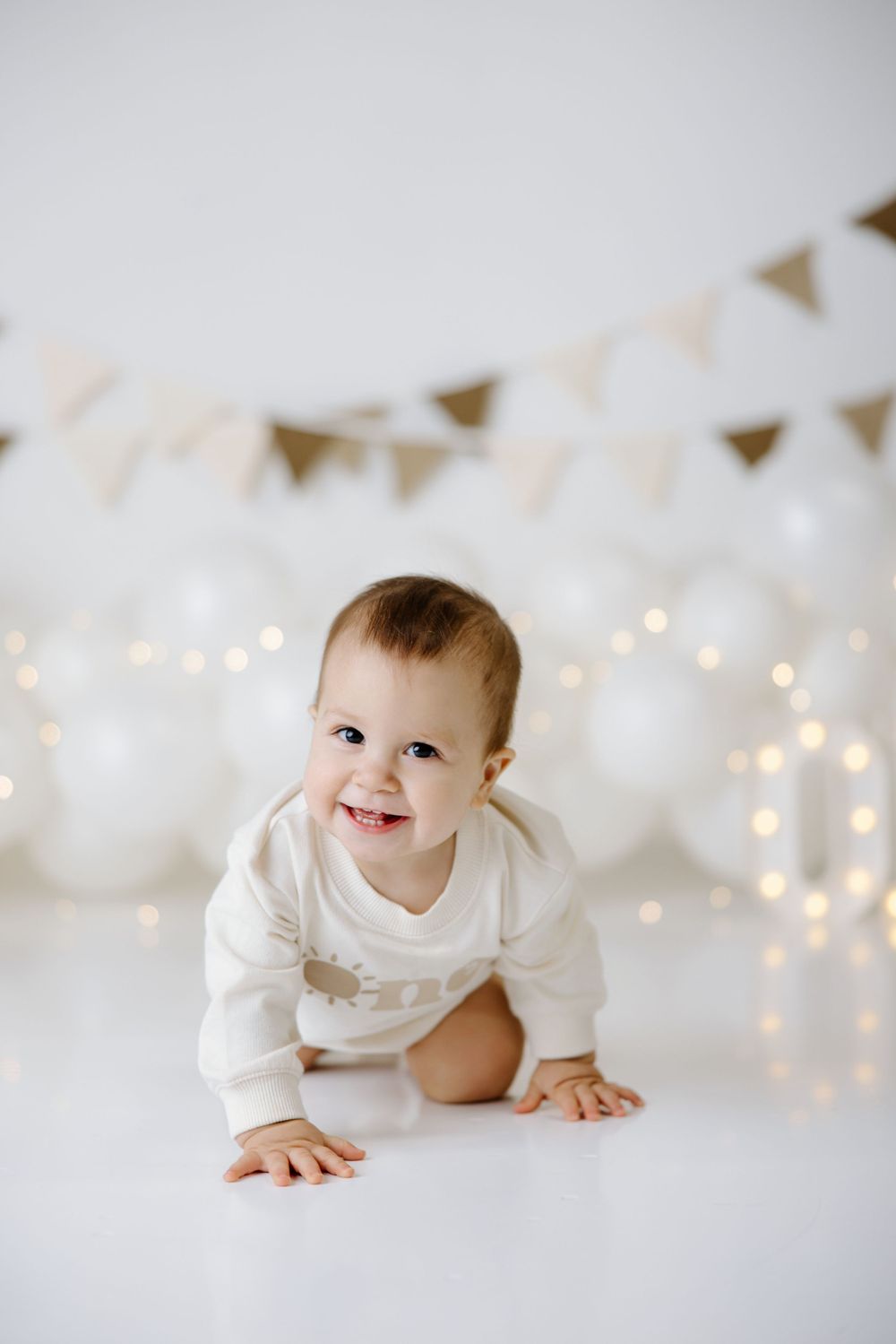 White and gold birthday photo setup with string lights and triangle bunting decorations.