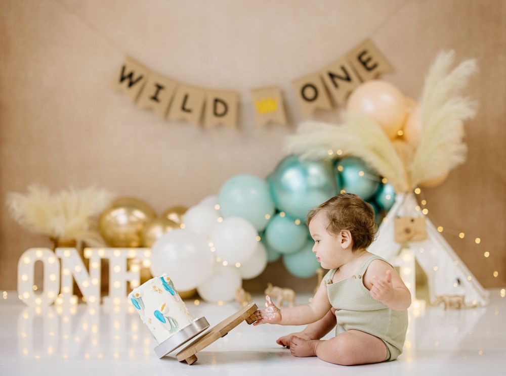 Baby crawling on floor with 'Wild One' banner and teal balloon arch in background.