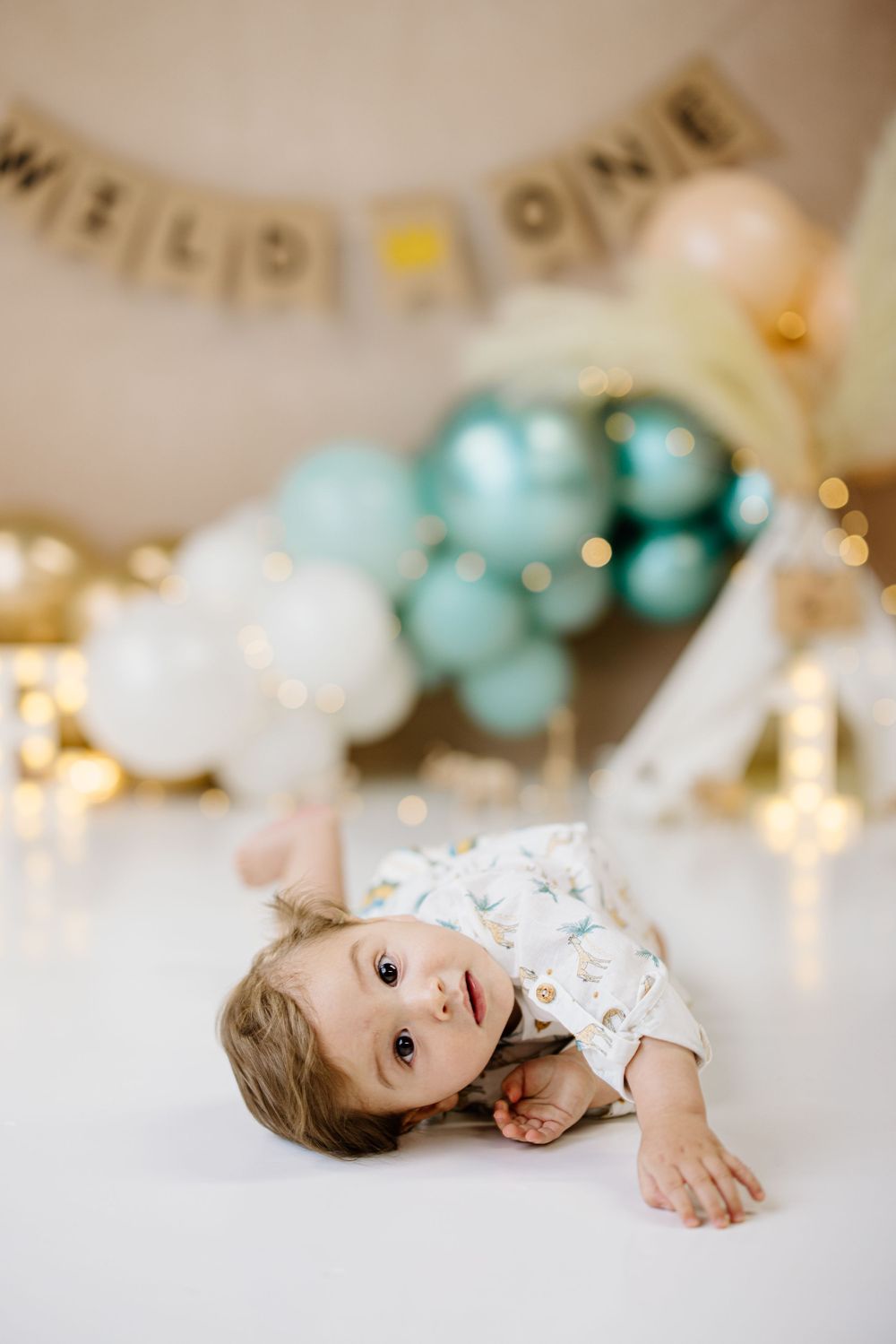 Baby laying on white floor with teal balloons and 'Wild One' banner in backdrop for first birthday photoshoot.
