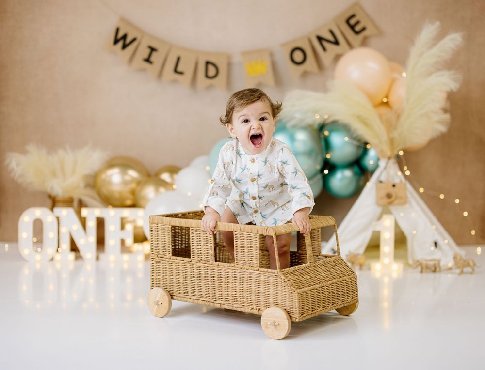 Baby sitting in woven toy car with festive balloon decor and teepee for first birthday celebration.