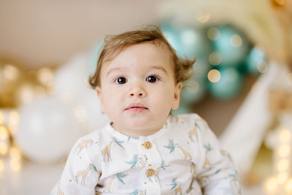Baby wearing white patterned outfit with teal balloons and gold decor in background.