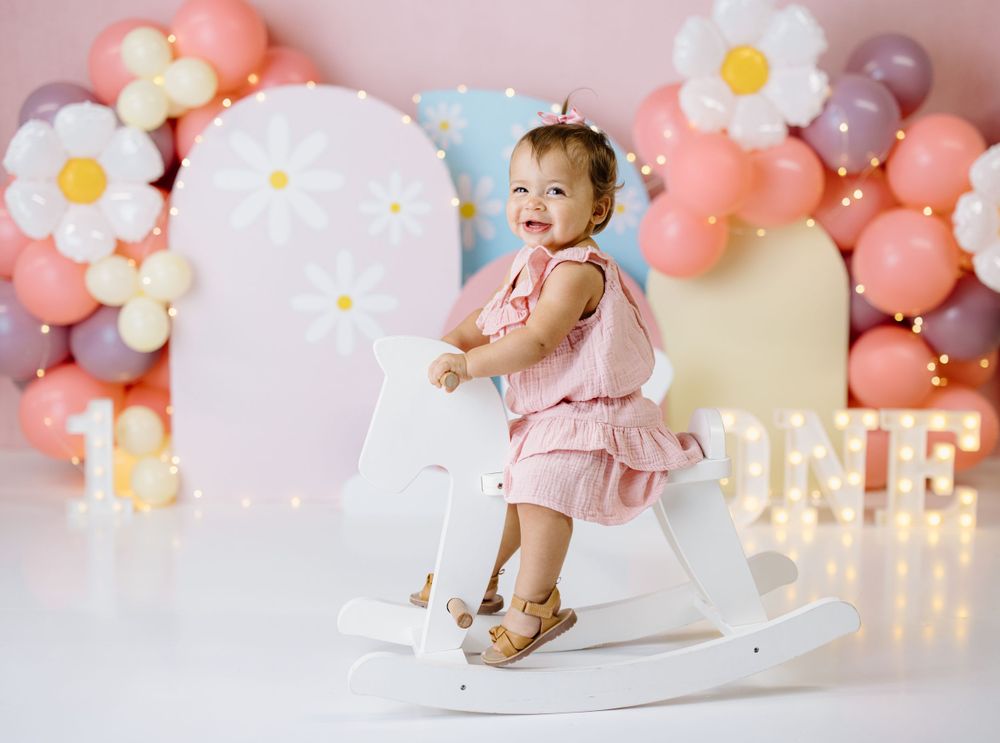 Baby on rocking horse with pink and white balloon arch and daisy decorations for birthday photoshoot.