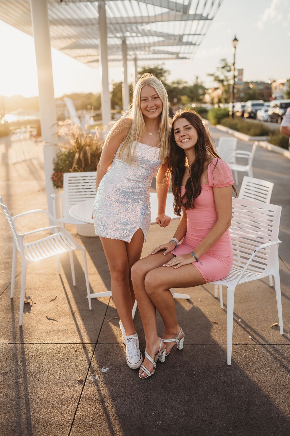 Two friends in dresses posing on white chairs on a patio during sunset.