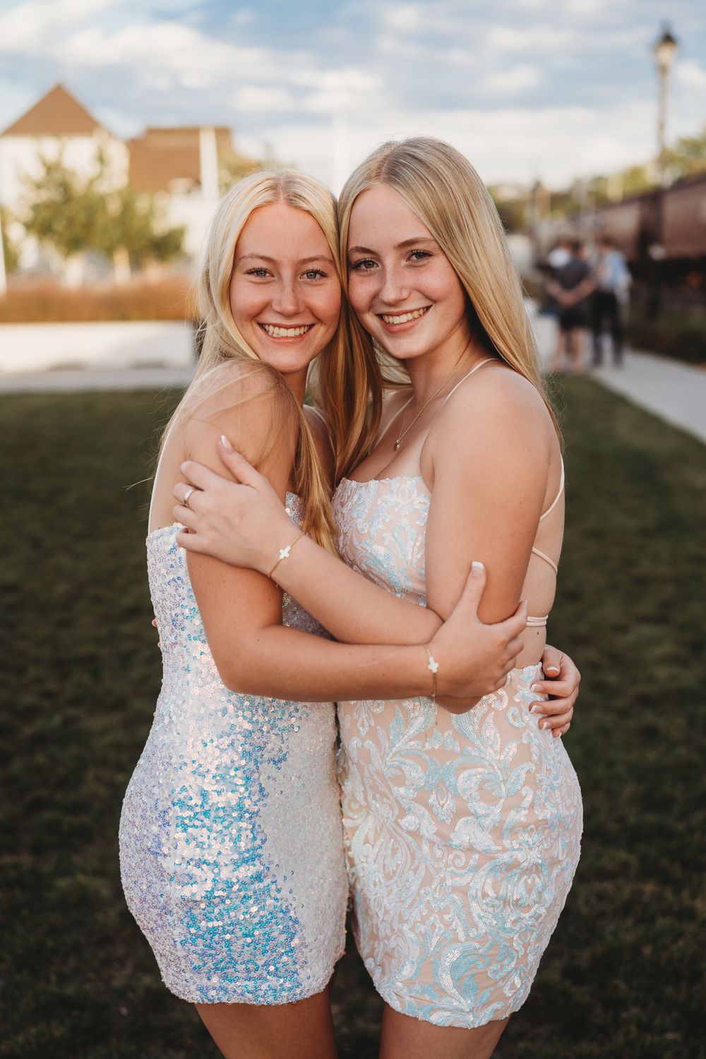 Two women in sparkly white dresses pose together at an outdoor event during golden hour.