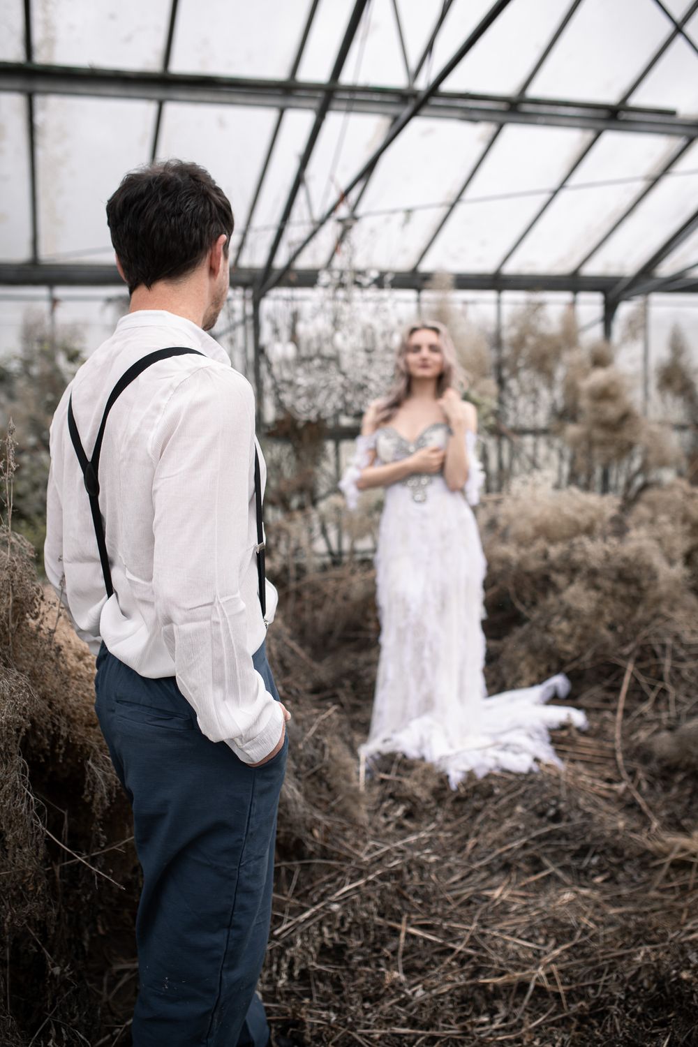 A couple in formal attire poses dramatically in a rustic greenhouse setting with dried brush and branches.