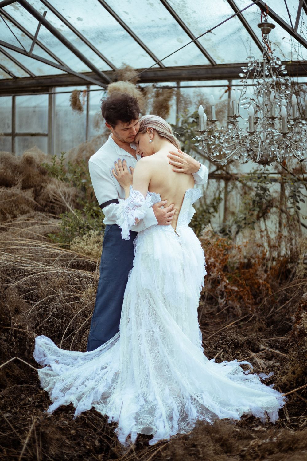 A romantic wedding couple embraces in a rustic greenhouse, with the bride wearing a flowing white lace gown.