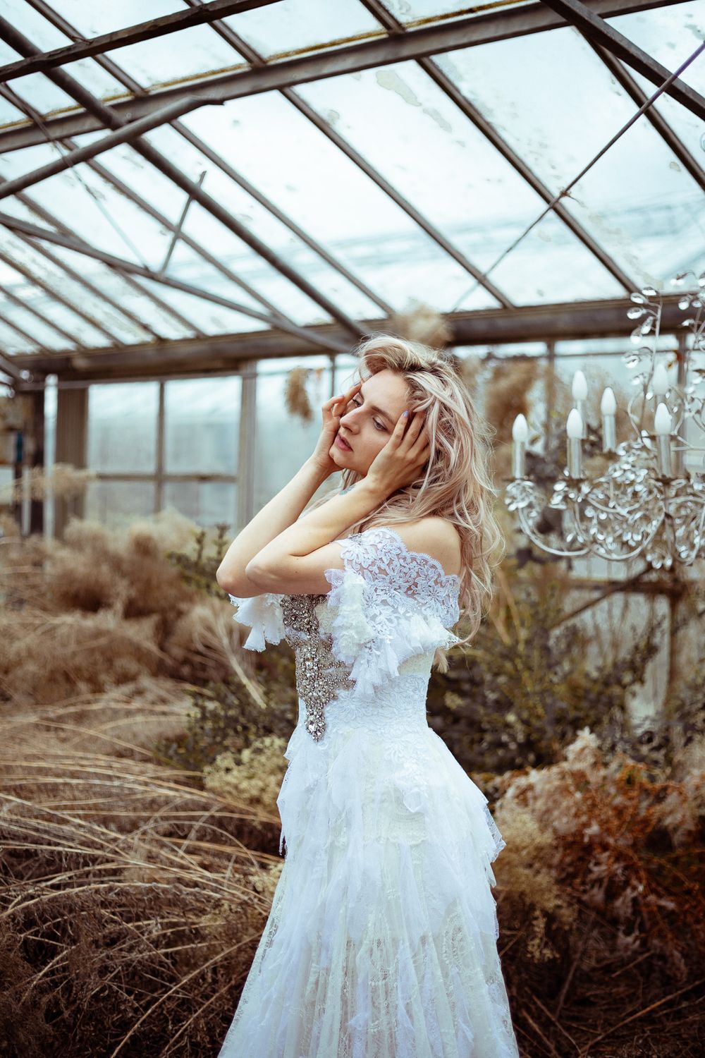 A dreamy greenhouse wedding scene features a flowing white lace gown among dried plants and crystal chandeliers.