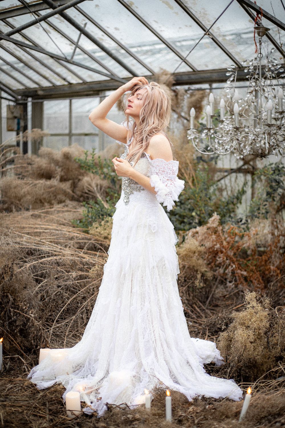 A figure in a flowing white lace dress twirls and poses gracefully inside a rustic greenhouse setting.