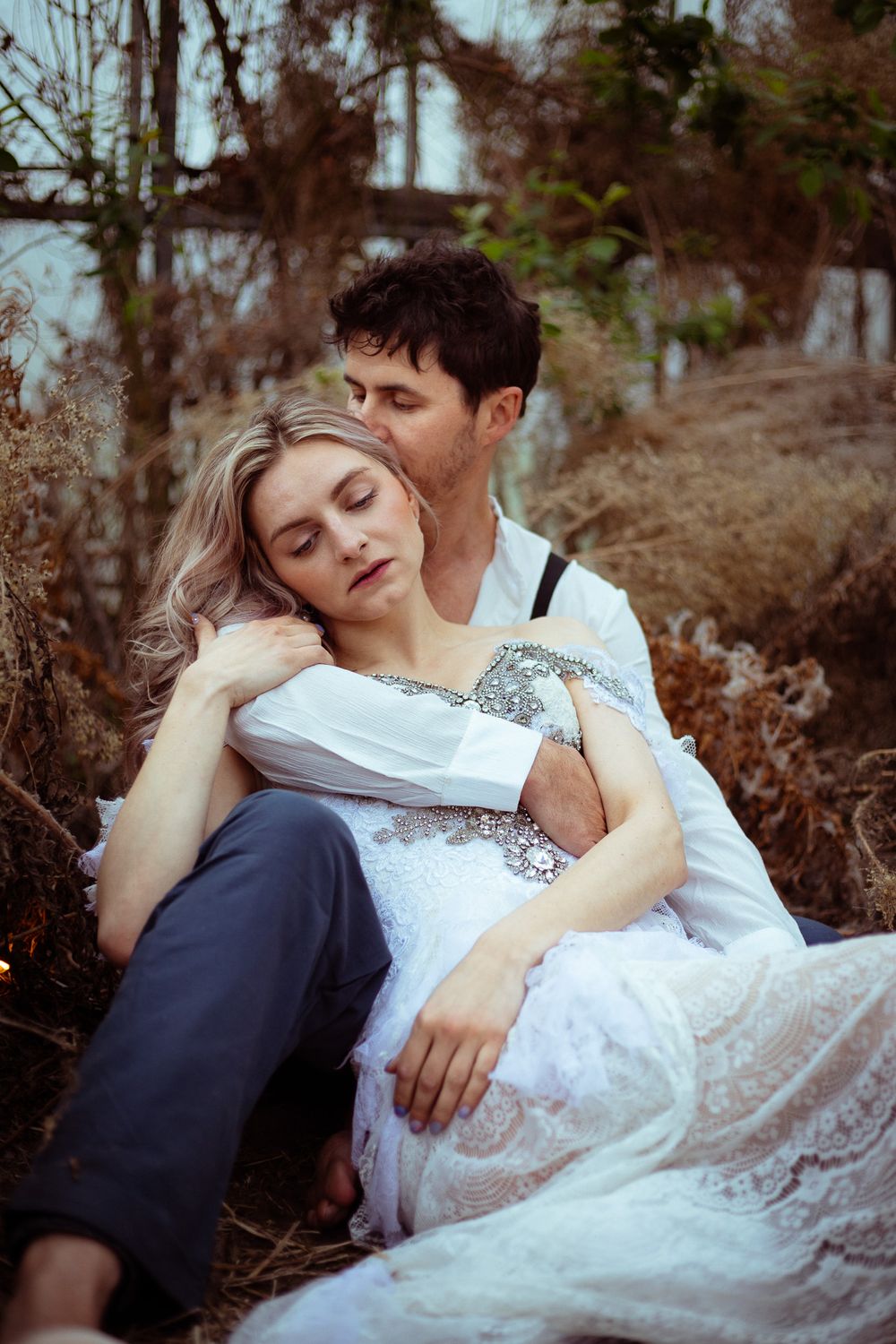 A couple in romantic embrace lies on autumn ground, with the person in white lace dress resting against their partner.