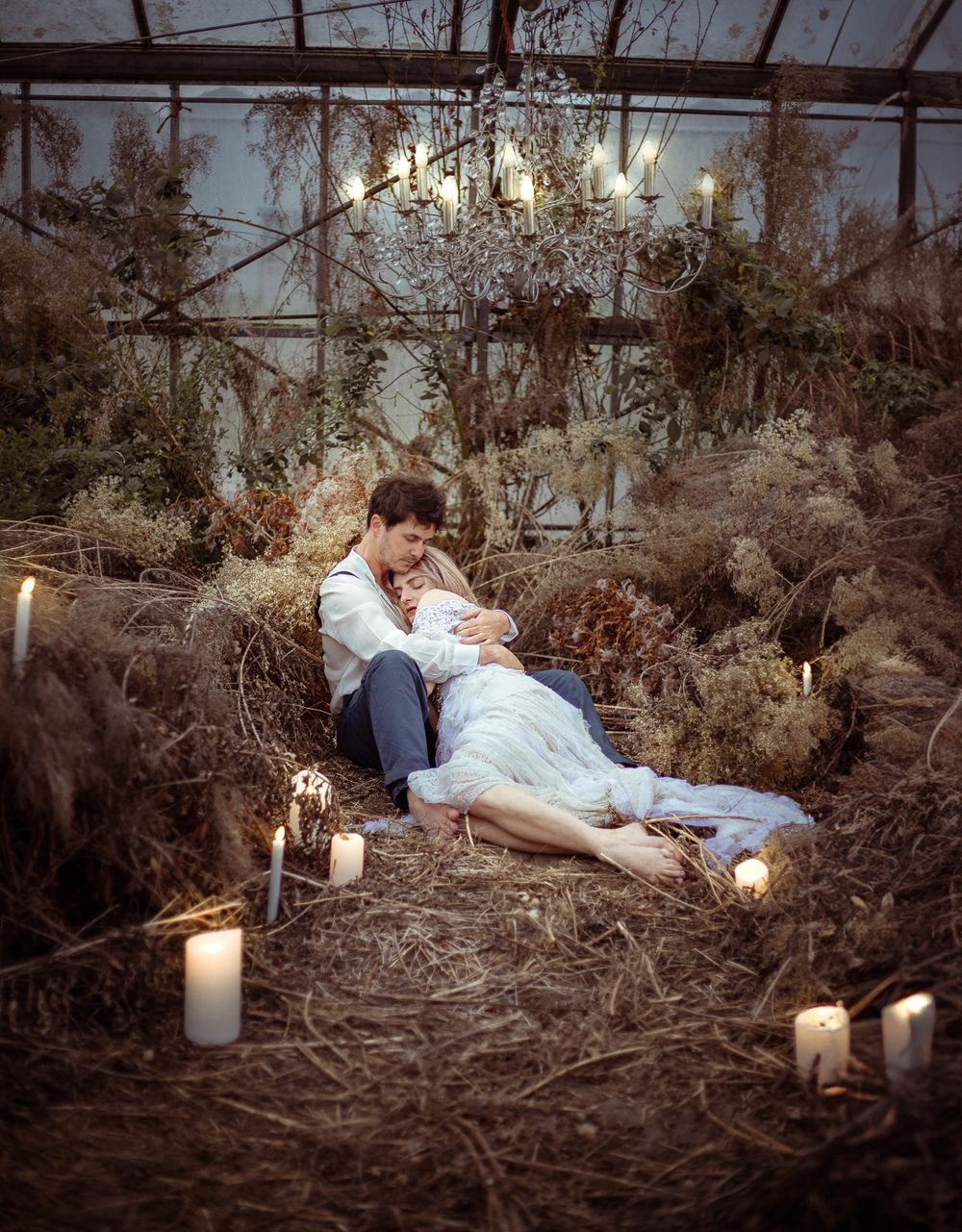 A romantic scene with lit candles and dried foliage in a rustic greenhouse setting creates a dreamy atmosphere.