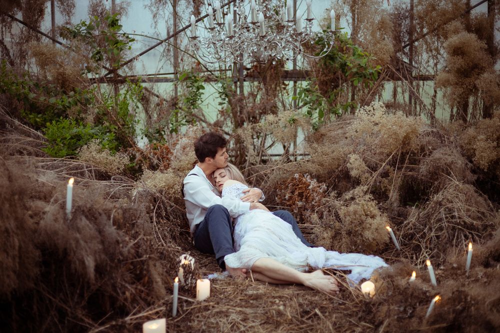 A couple in wedding attire poses together among dry grass in a dramatic greenhouse setting.