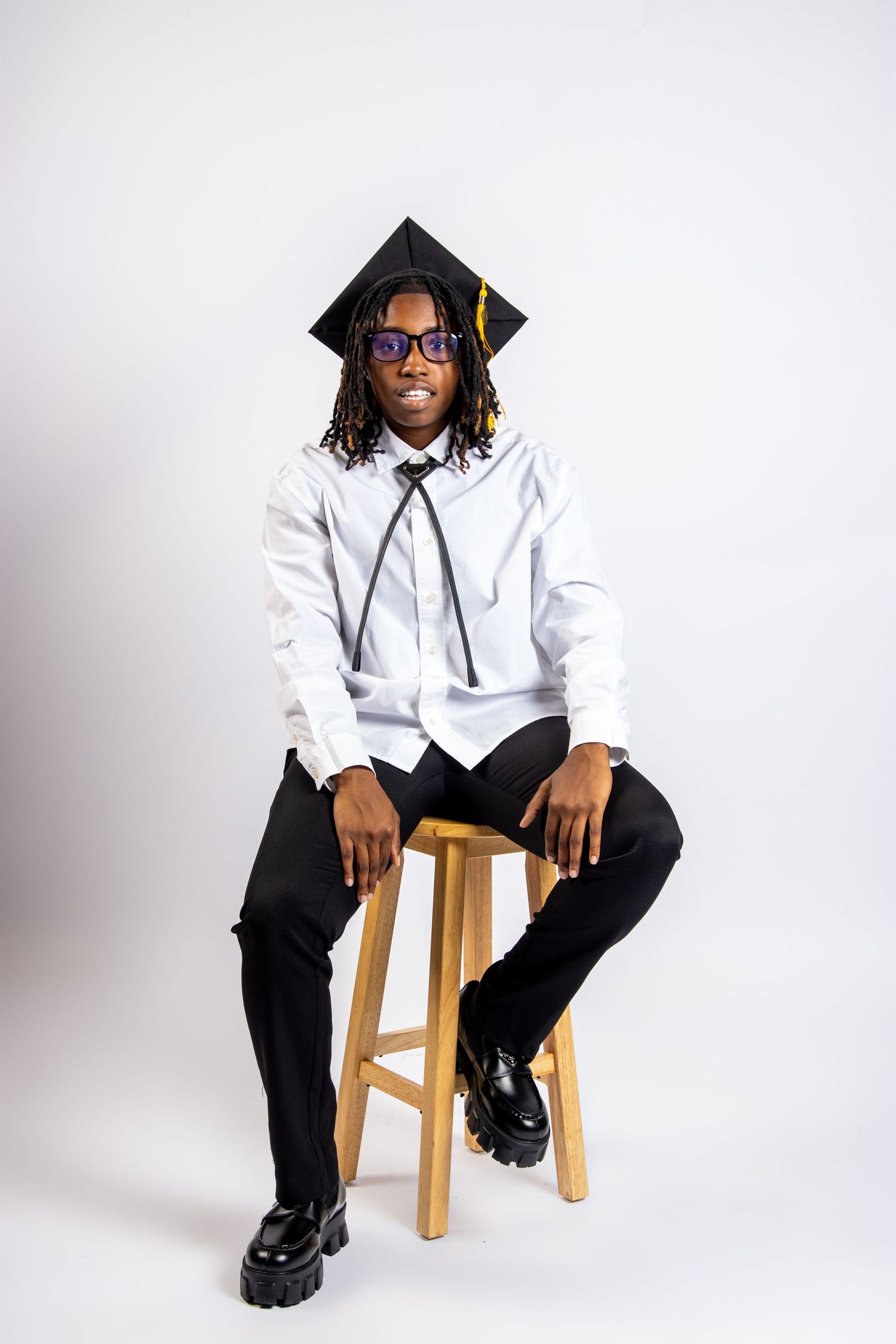 Smiling young man in graduation cap, white shirt, and black pants sitting on a wooden stool against white background.