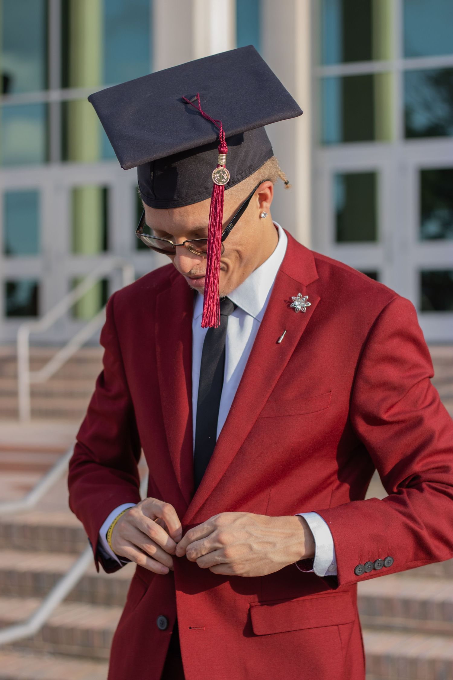 A graduate in a red suit and mortarboard cap buttons his jacket on university steps.