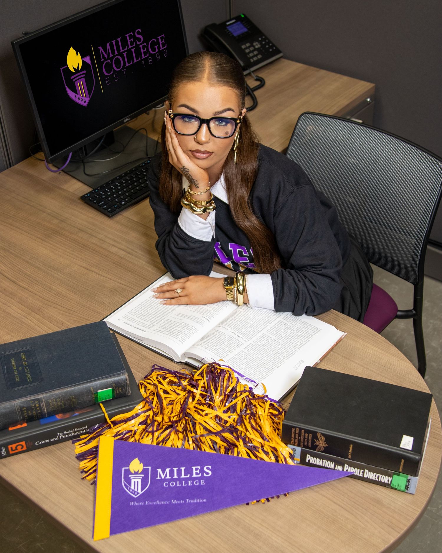 Woman in academic attire sitting at a desk with Miles College pennant, books, and cheerleading pom-poms.