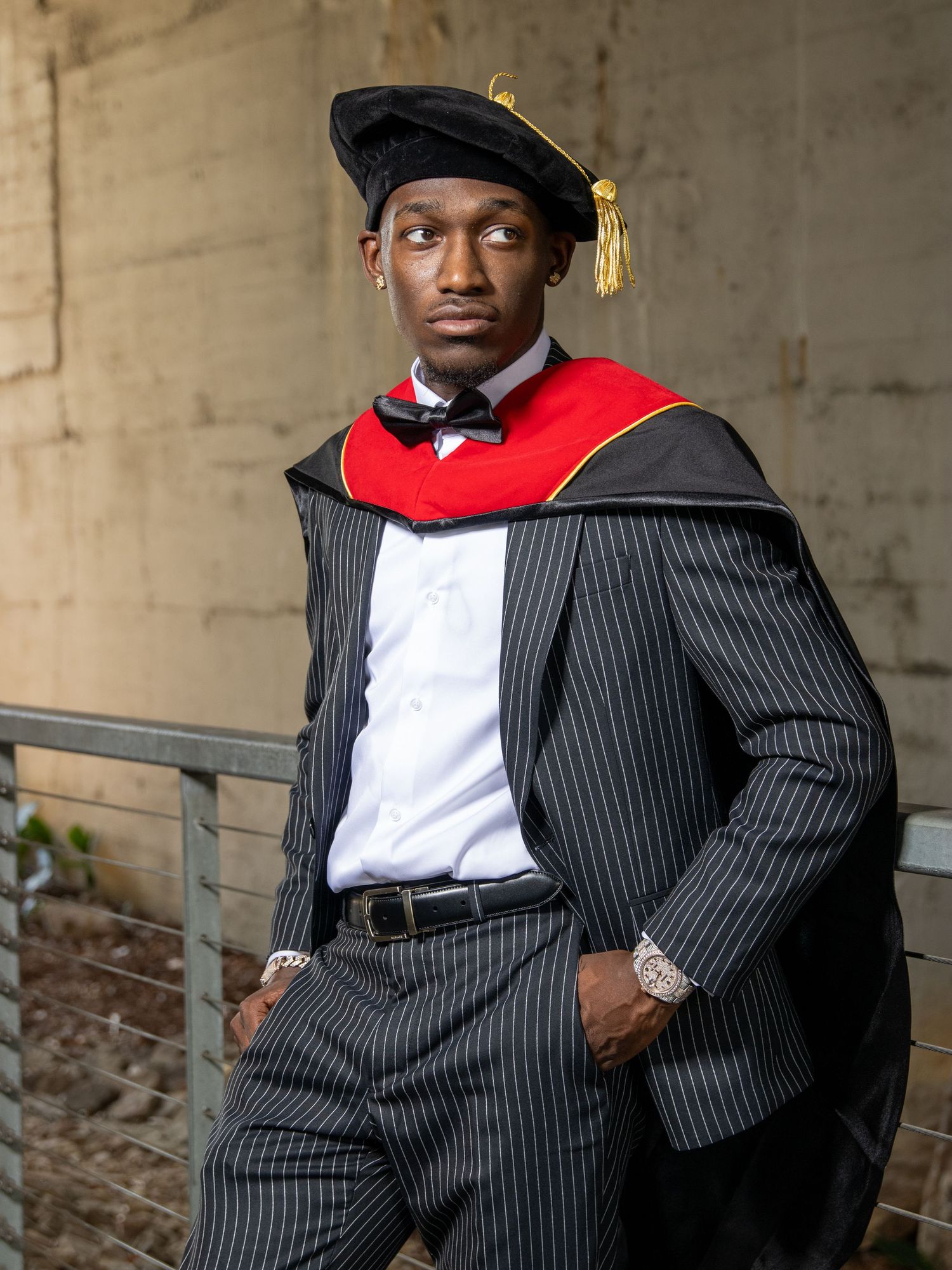Young man in doctoral graduation regalia with pinstripe suit posing confidently outdoors.