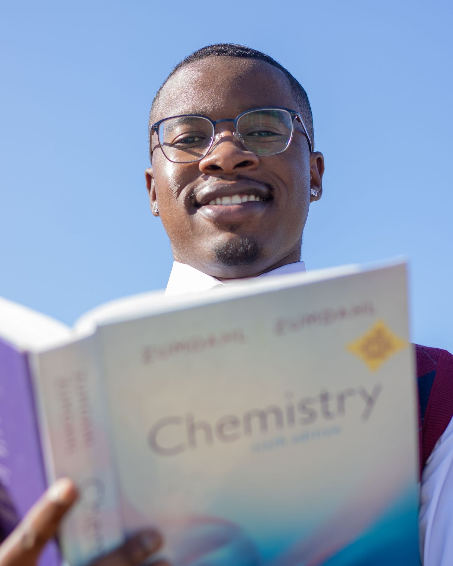 Smiling graduate holding Chemistry textbook outdoors in graduation attire under blue sky.