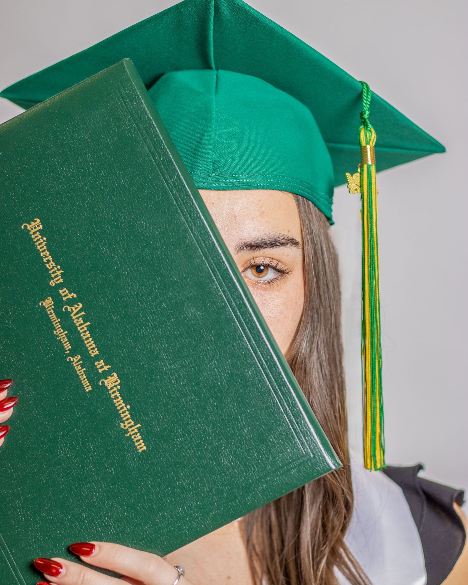 UAB graduate holding green diploma cover near face wearing cap and gown.