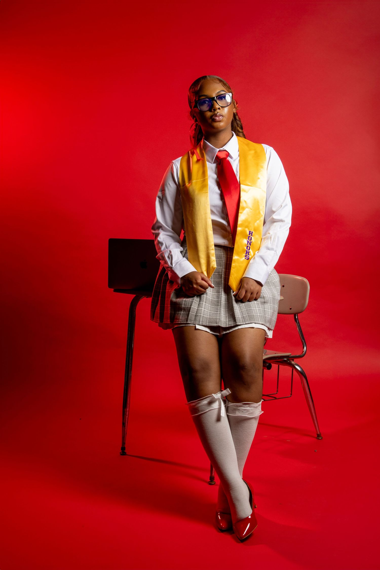Student in school uniform with yellow vest and tie poses against red background with vintage classroom chair.