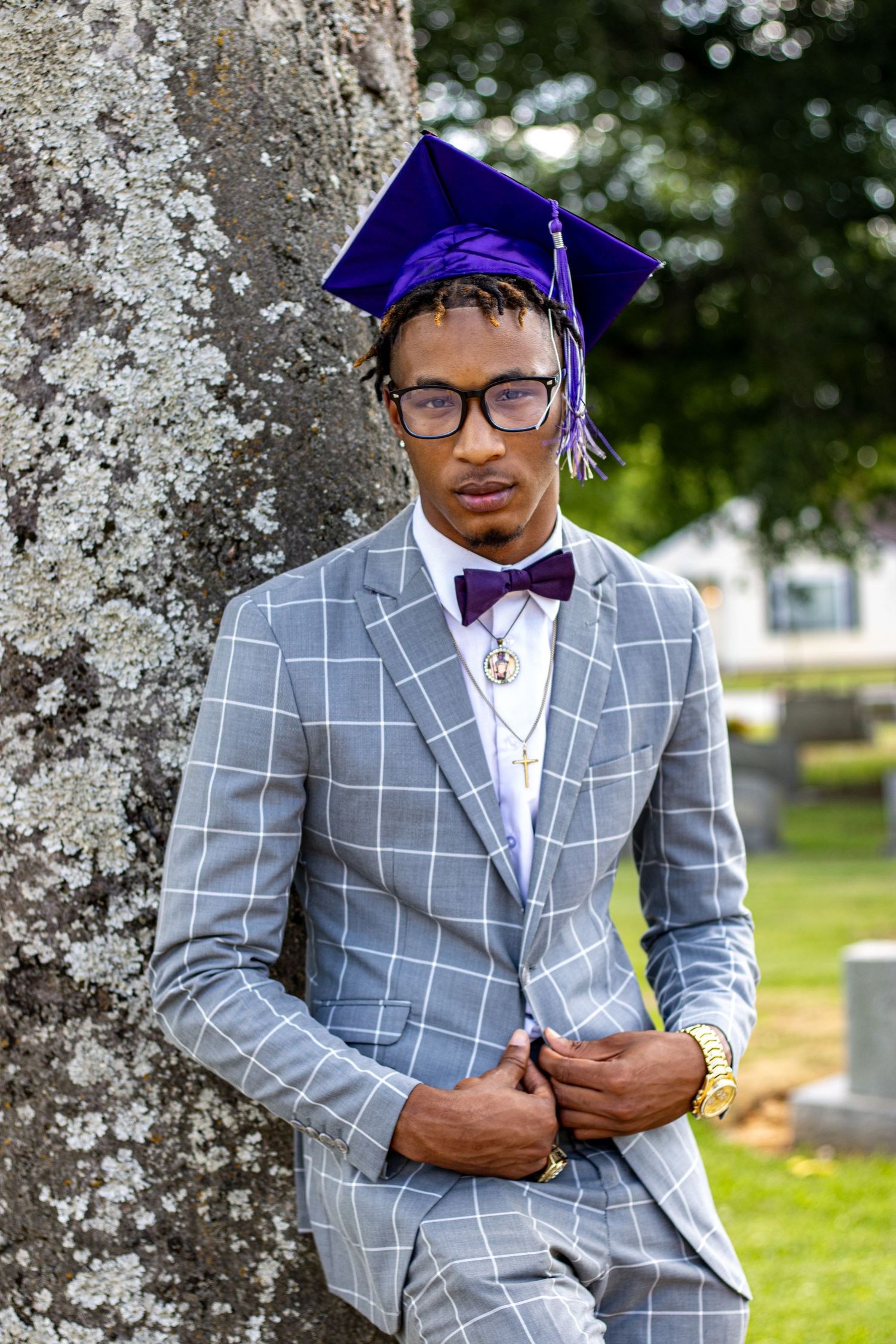 Young man in gray plaid suit and purple graduation cap poses against a moss-covered tree outdoors.