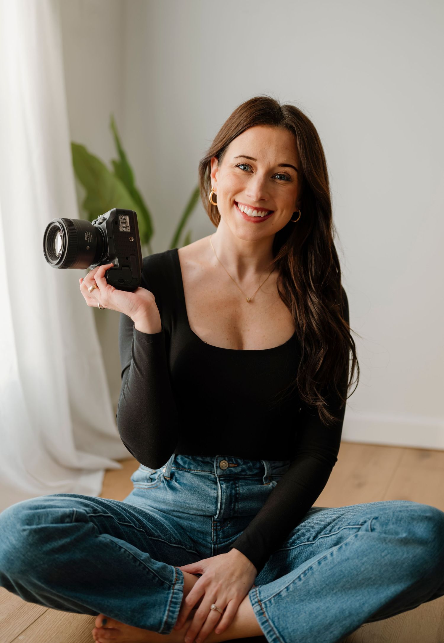 A photographer sits cross-legged on the floor holding a professional DSLR camera while wearing jeans and a black top.