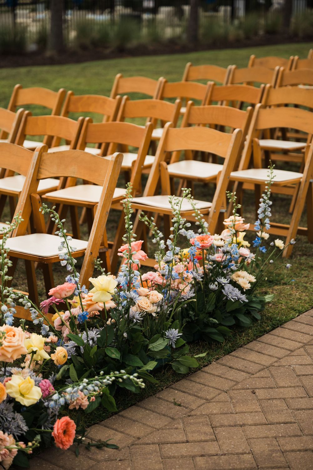 Elegant wedding ceremony setup with wooden chairs and colorful floral aisle arrangements on a brick pathway.