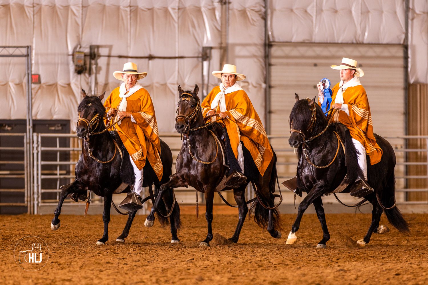 Three riders in orange capes on black horses perform a synchronized equestrian routine indoors on a dirt arena.