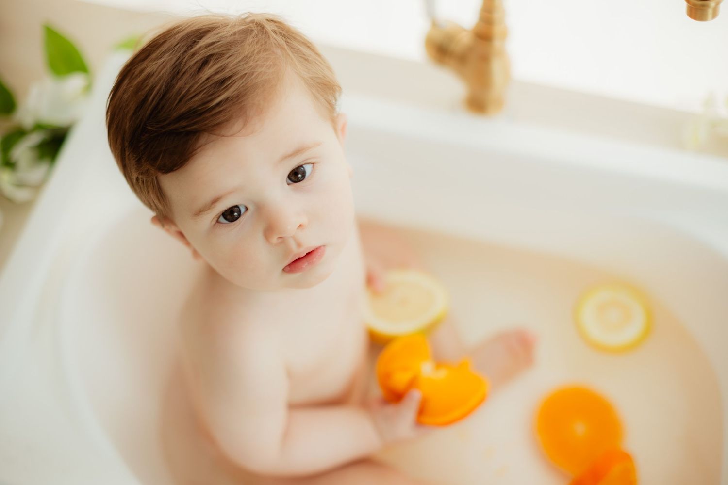 Cute toddler sitting in a bathtub with orange and lemon citrus fruit slices, looking up at the camera.