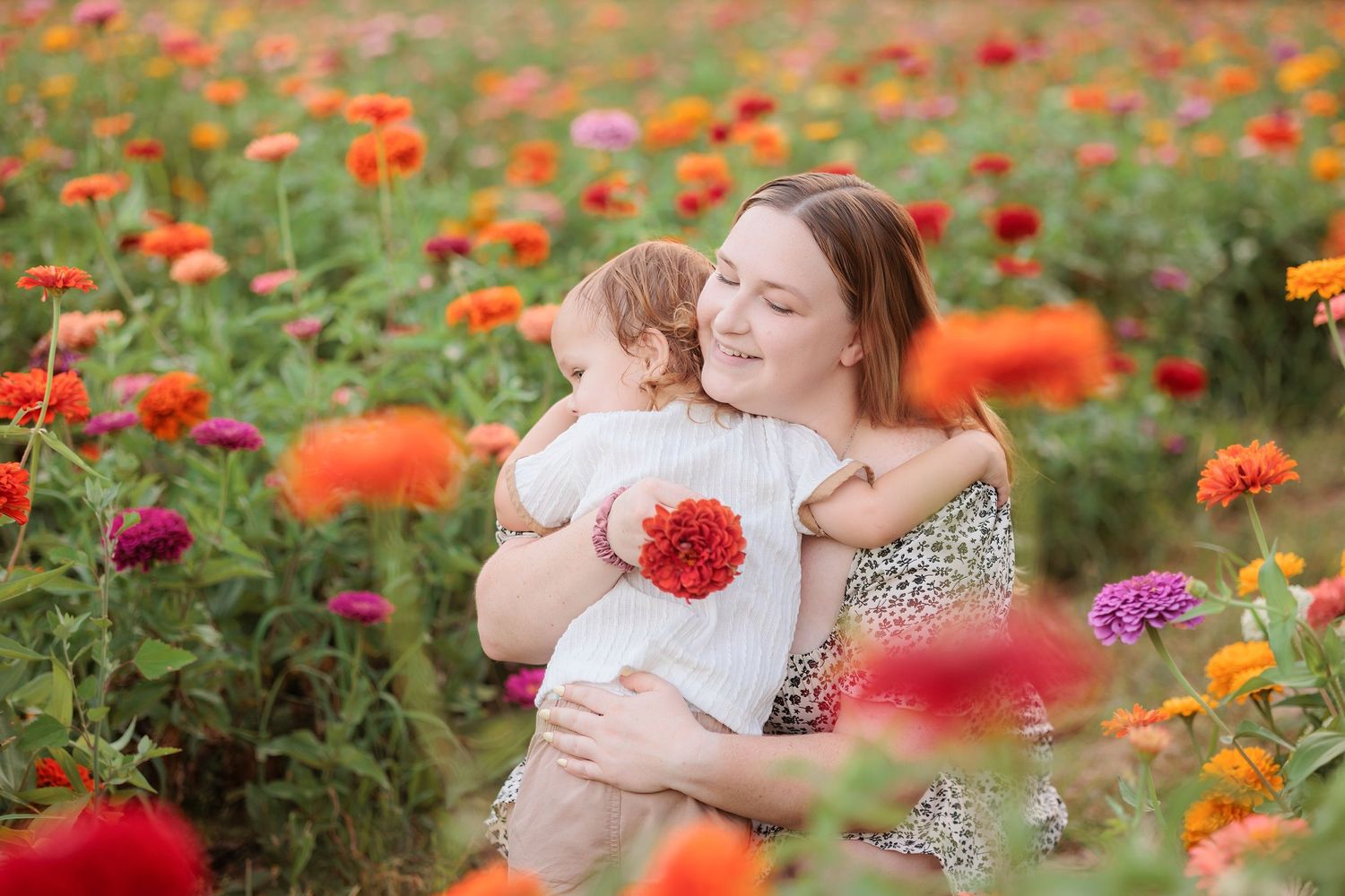 Mother holding baby girl surrounded by colorful zinnia flowers in a vibrant garden field.