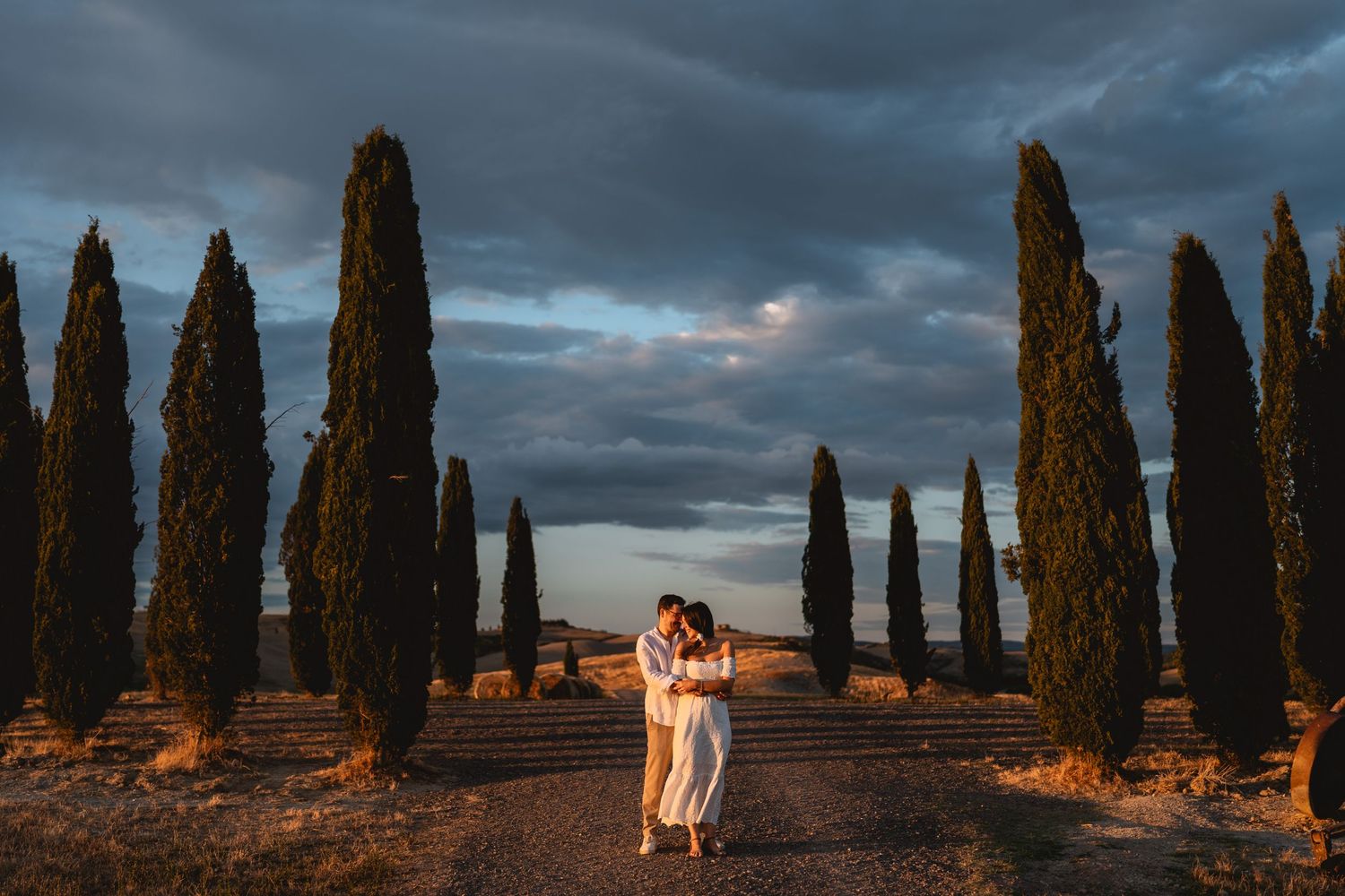 Couple embracing at sunset on a cypress-lined Tuscan road under dramatic stormy skies.