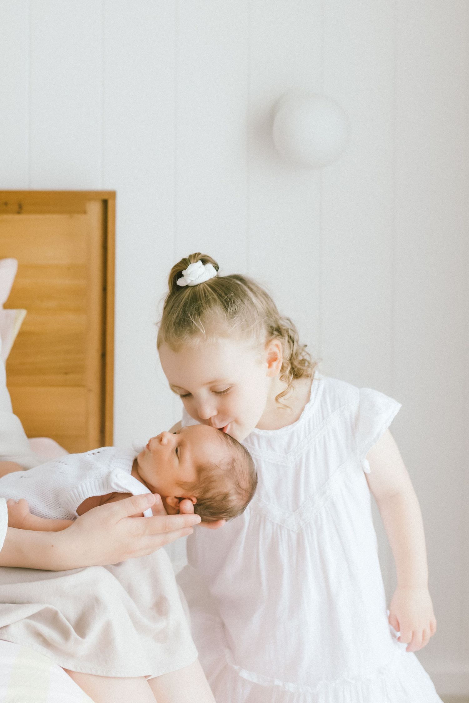 A young girl in a white dress gently kisses a newborn baby held by an adult in a bright, airy room.