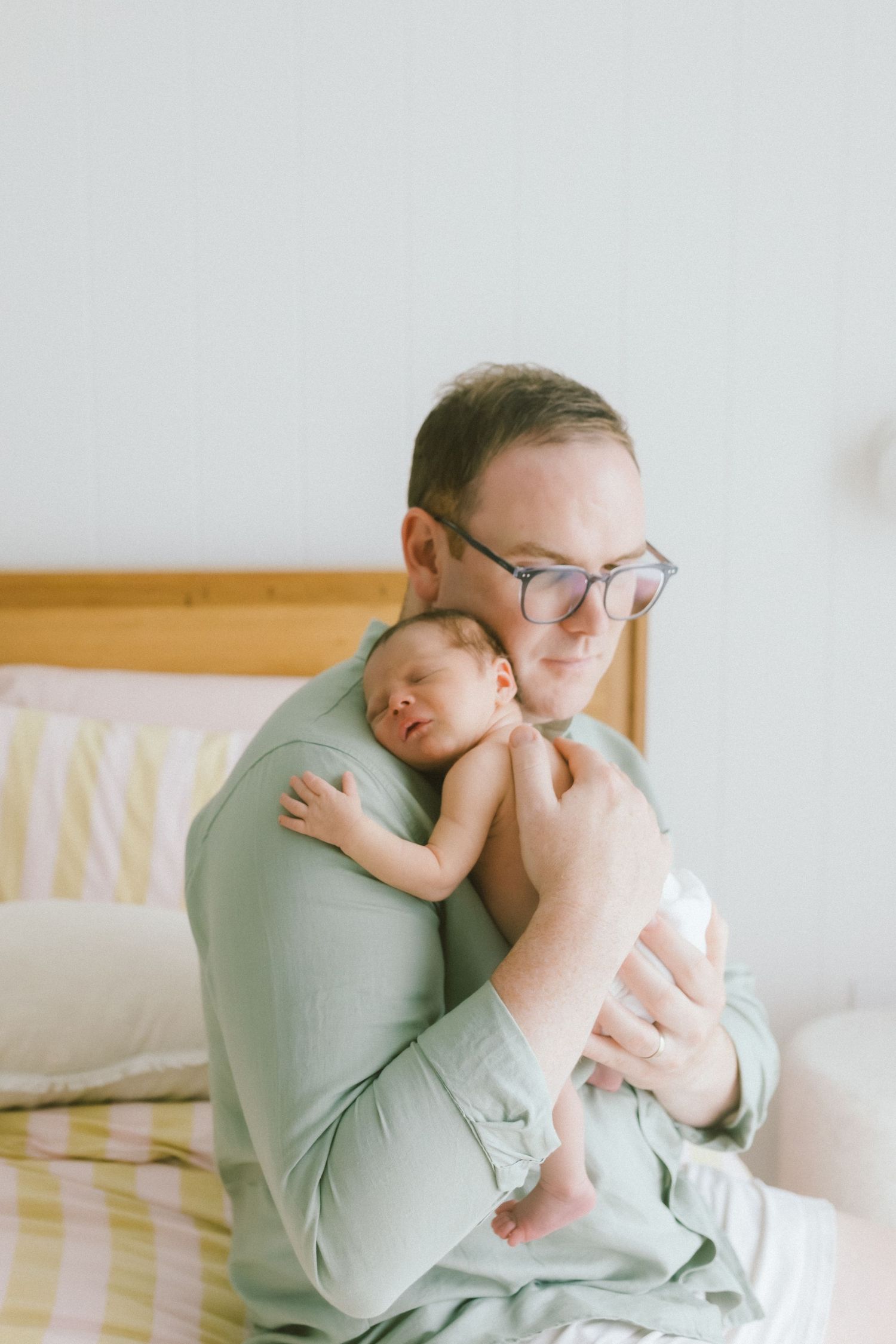 Father in green shirt holding sleeping newborn baby close to his chest while sitting near a bed.