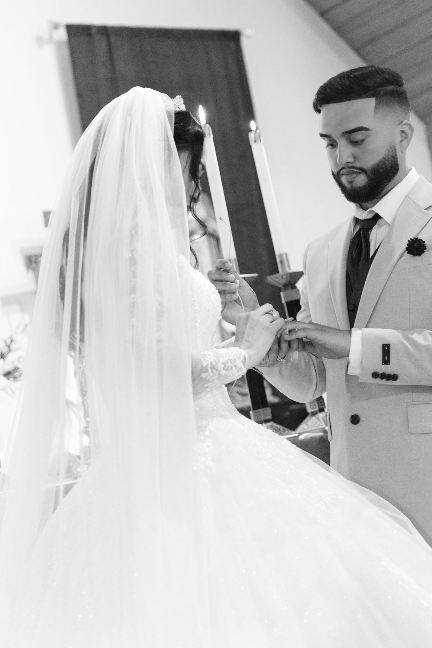 Groom placing wedding ring on bride's finger at church altar with candles and red curtain backdrop.