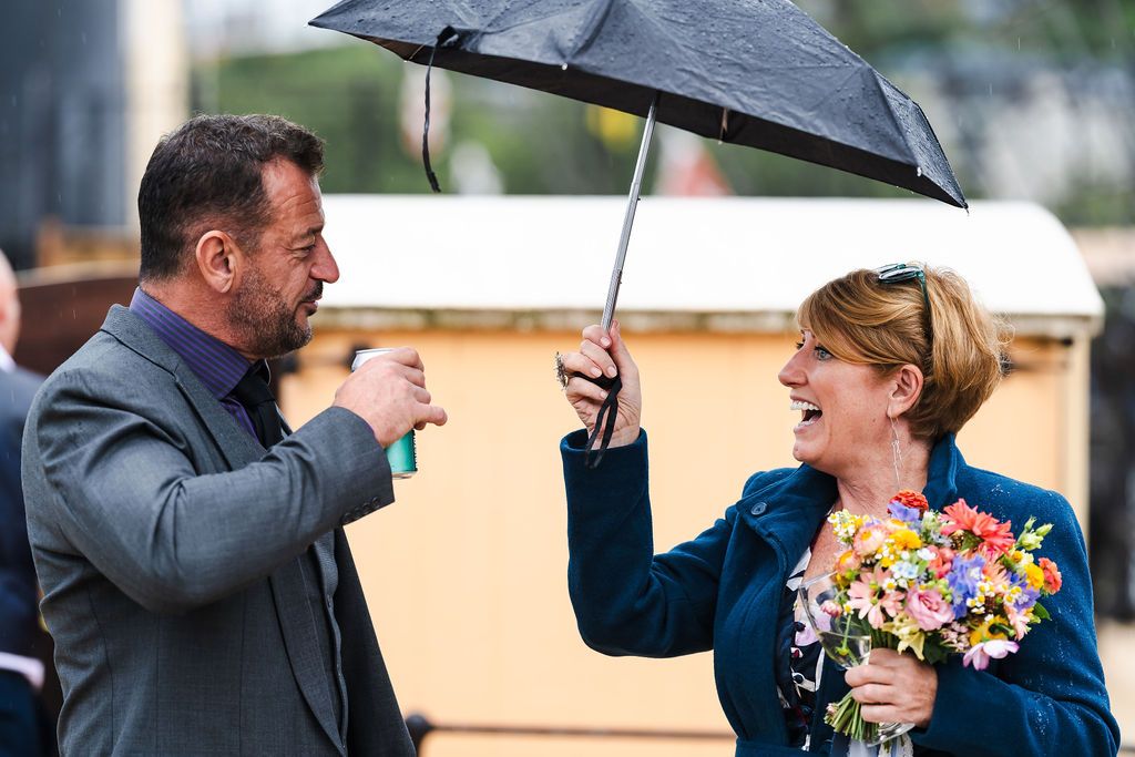 Man and woman sharing umbrella in rain, woman holding colorful flower bouquet, smiling outdoors.